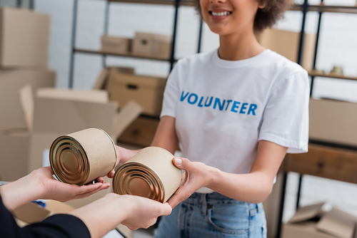 cropped view of smiling african american volunteer handing out canned food in charity center