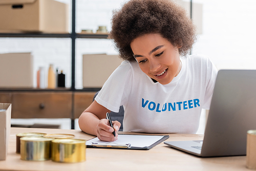 smiling african american volunteer writing on clipboard near blurred laptop