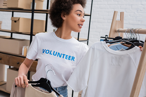 smiling african american volunteer holding hangers with clothes in charity storehouse