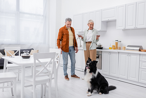 Cheerful woman holding plates near husband and border collie in kitchen