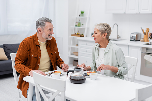 Cheerful mature man talking to wife near tasty pancakes and coffee in kitchen