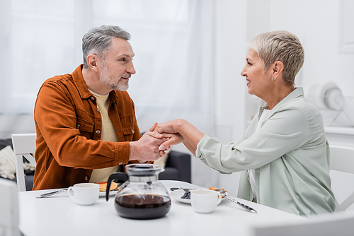 Positive couple holding hands near coffee and pancakes in kitchen