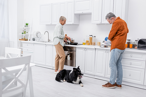 Side view of man pouring dog food in bowl near border collie and wife cooking in kitchen