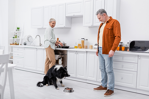 Smiling couple looking at border collie sitting near bowl with food in kitchen