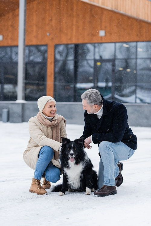 Happy senior woman in winter outfit looking at husband near border collie on snow outdoors