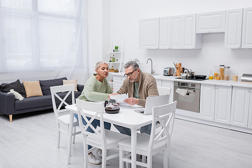 Senior woman talking to husband holding paper near coffee and laptop in kitchen