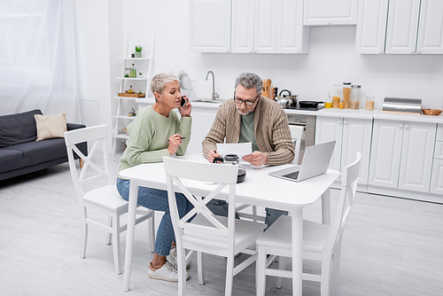 Senior woman talking on smartphone while husband holding paper in kitchen
