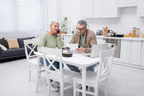 Elderly woman talking on smartphone while husband holding paper near coffee and laptop in kitchen