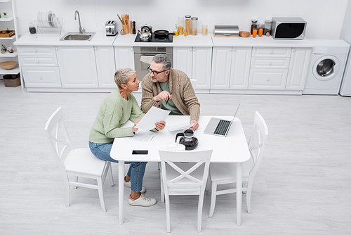 High angle view of pensioners holding papers and talking near coffee and gadgets in kitchen