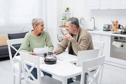 Cheerful senior couple talking near papers and laptop in kitchen