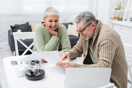 Positive pensioner pointing at paper near wife and devices in kitchen