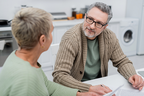Mature man looking at blurred wife near documents in kitchen