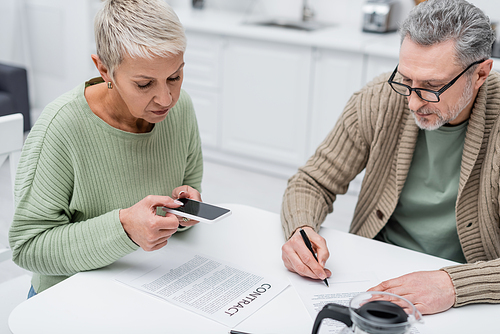 Elderly woman taking photo of contract near husband with document and coffee in kitchen