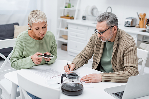Pensioner writing on document near wife taking photo of contract in kitchen