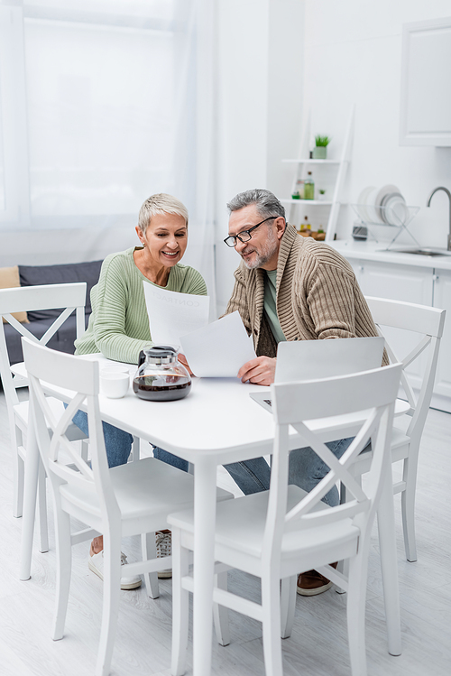 Cheerful senior woman holding paper near husband, laptop and coffee in kitchen