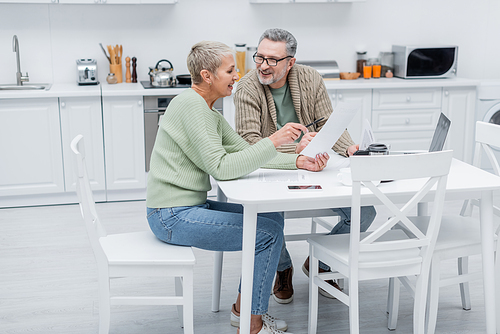 Positive pensioners holding documents near devices in kitchen