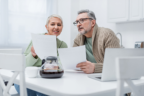 Positive elderly woman holding document near husband, laptop and blurred coffee in kitchen