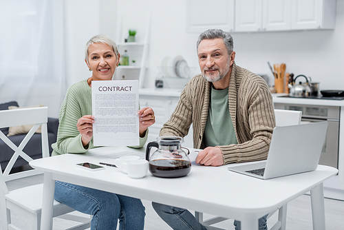 Pensioners holding contract and looking at camera near devices and coffee in kitchen
