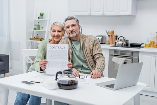 Smiling senior woman holding contract near husband and devices in kitchen