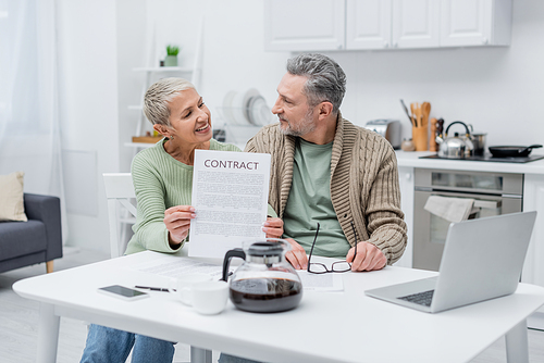Cheerful senior woman holding contract and looking at husband near gadgets and coffee in kitchen