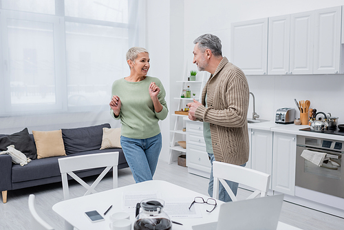 Cheerful senior woman dancing near husband, papers and devices in kitchen