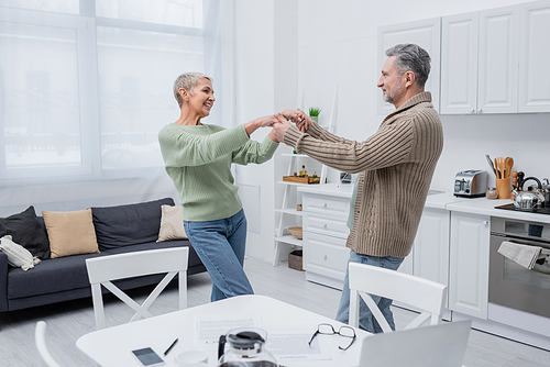 Positive couple dancing near papers and devices in kitchen