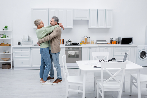 Side view of senior woman hugging husband near papers and devices in kitchen