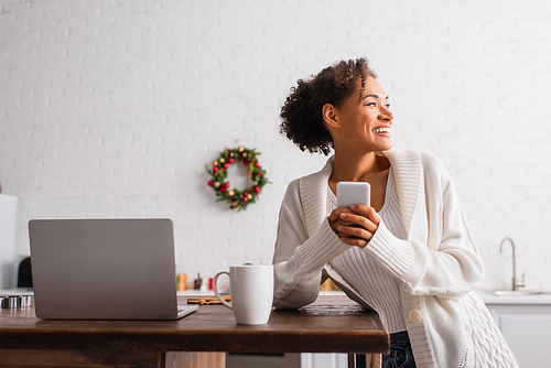 Positive african american woman using smartphone near cup and laptop in kitchen on christmas