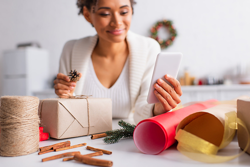 Smartphone in hand of blurred african american woman decorating christmas gift at home