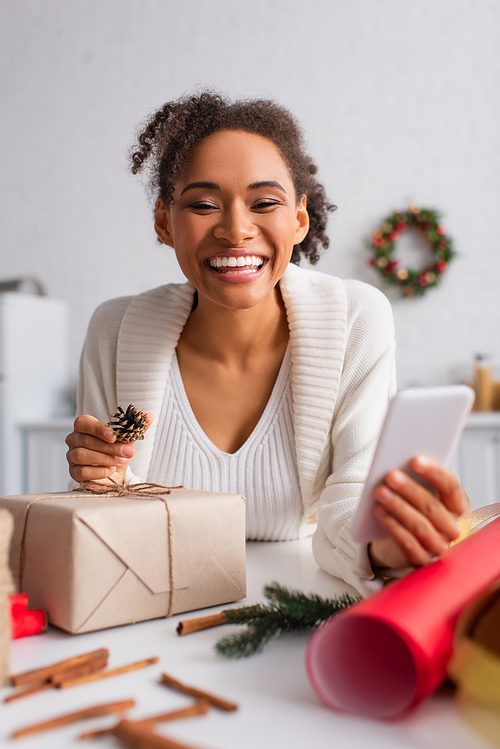 Cheerful african american woman holding smartphone while decorating present at home