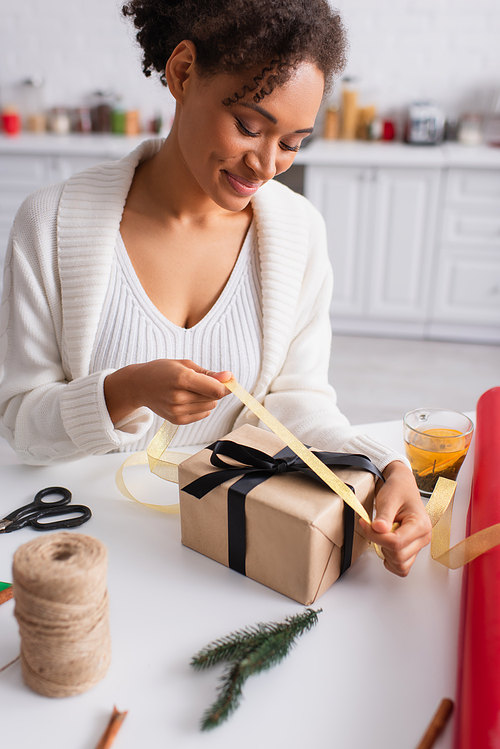 Happy african american woman holding ribbon while decorating christmas gift at home