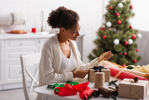 African american woman decorating christmas gift with ribbon at home