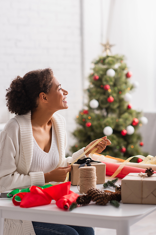 Side view of cheerful african american woman decorating present near blurred christmas tree