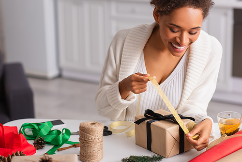 Positive african american woman holding ribbon while decorating present near cup of tea at home