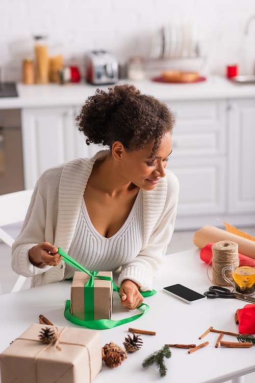 African american woman looking at smartphone while decorating present near tea at home