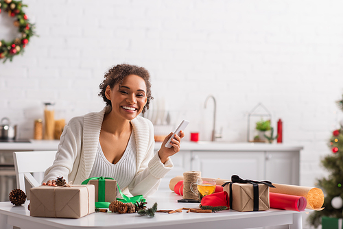Positive african american woman holding cellphone near presents, christmas decor and tea