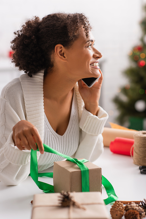 Side view of smiling african american woman talking on smartphone while wrapping christmas gifts