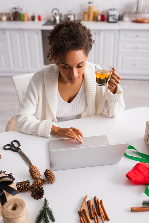 African american woman holding tea and using laptop near christmas decor at home