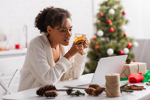 African american woman holding tea near laptop, present and christmas decor