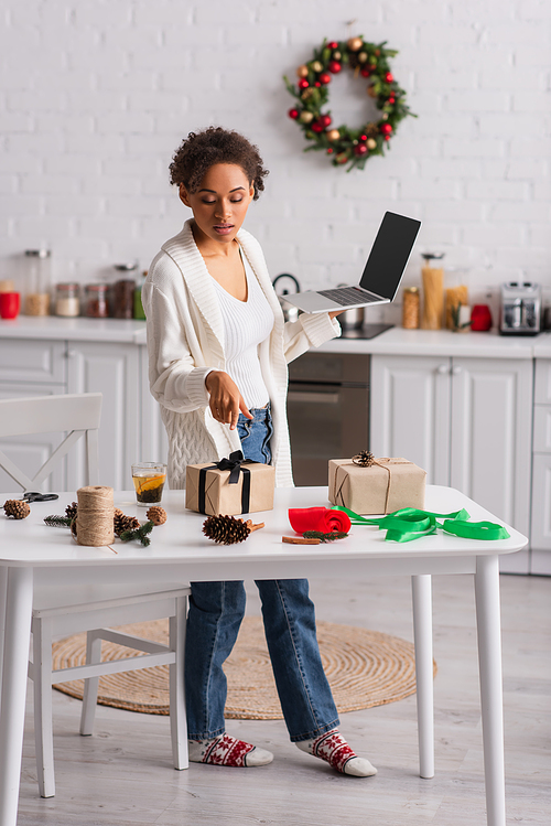 African american woman with laptop pointing at gift near christmas decor at home