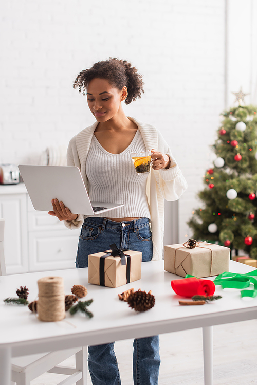 Young african american woman holding laptop and tea near gifts and christmas decor at home