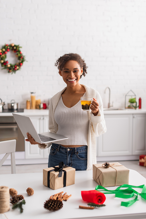 Smiling african american woman holding tea and laptop near gift boxes and christmas decor