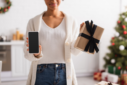 Cropped view of blurred african american woman holding smartphone and christmas gift at home