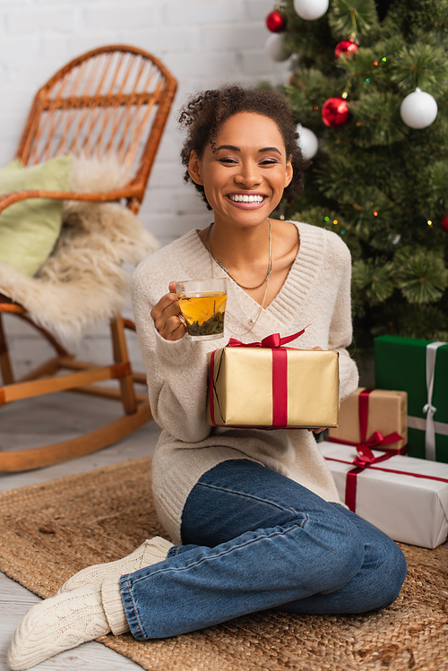 Positive african american woman holding gift and cup of tea near christmas tree at home