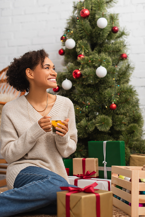 Cheerful african american woman holding tea near presents and christmas tree