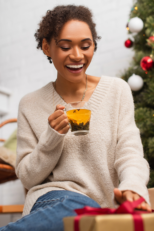 Happy african american woman with cup of tea looking at blurred christmas gift at home