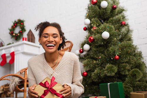 Positive african american woman holding present and looking at camera near blurred christmas tree at home