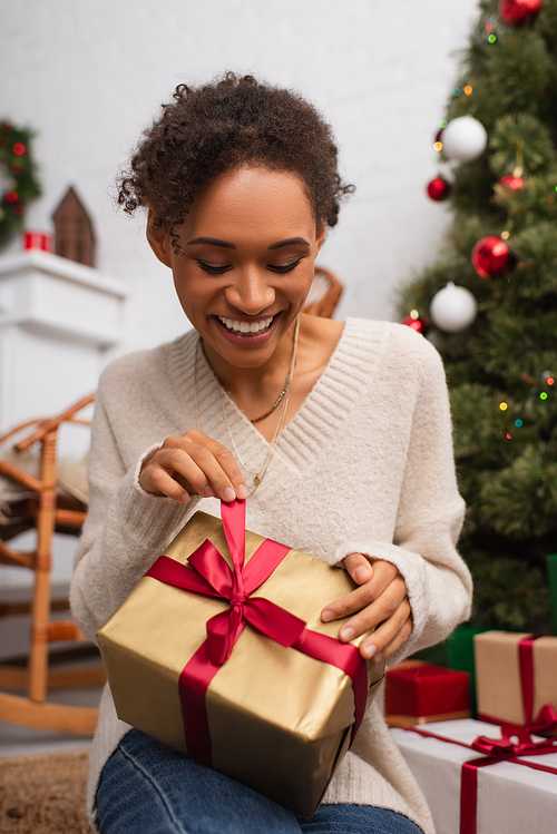 Young african american woman smiling while untying christmas gift at home