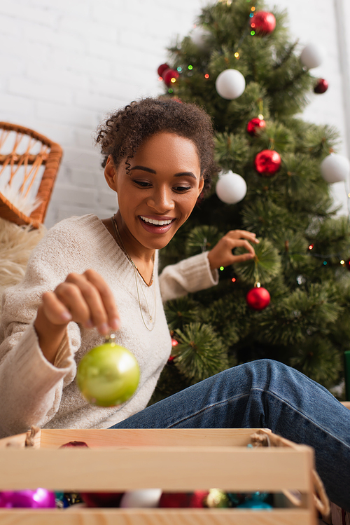 Happy african american woman holding ball near box and christmas tree at home