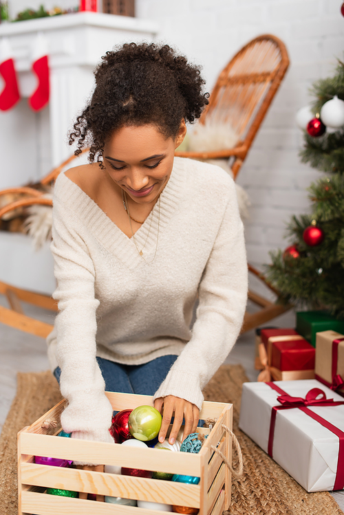 African american woman looking at box with christmas balls near gifts at home